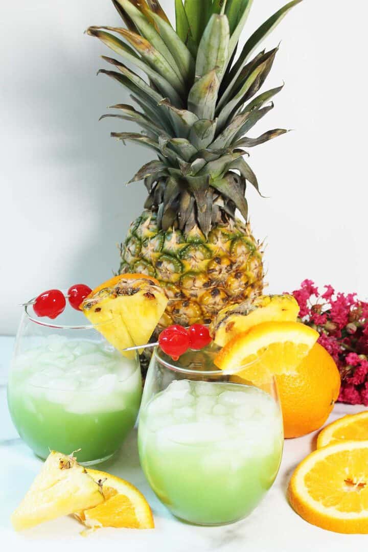 Two blue iguana cocktails sitting in front of pineapple with oranges and pineapple pieces on table.