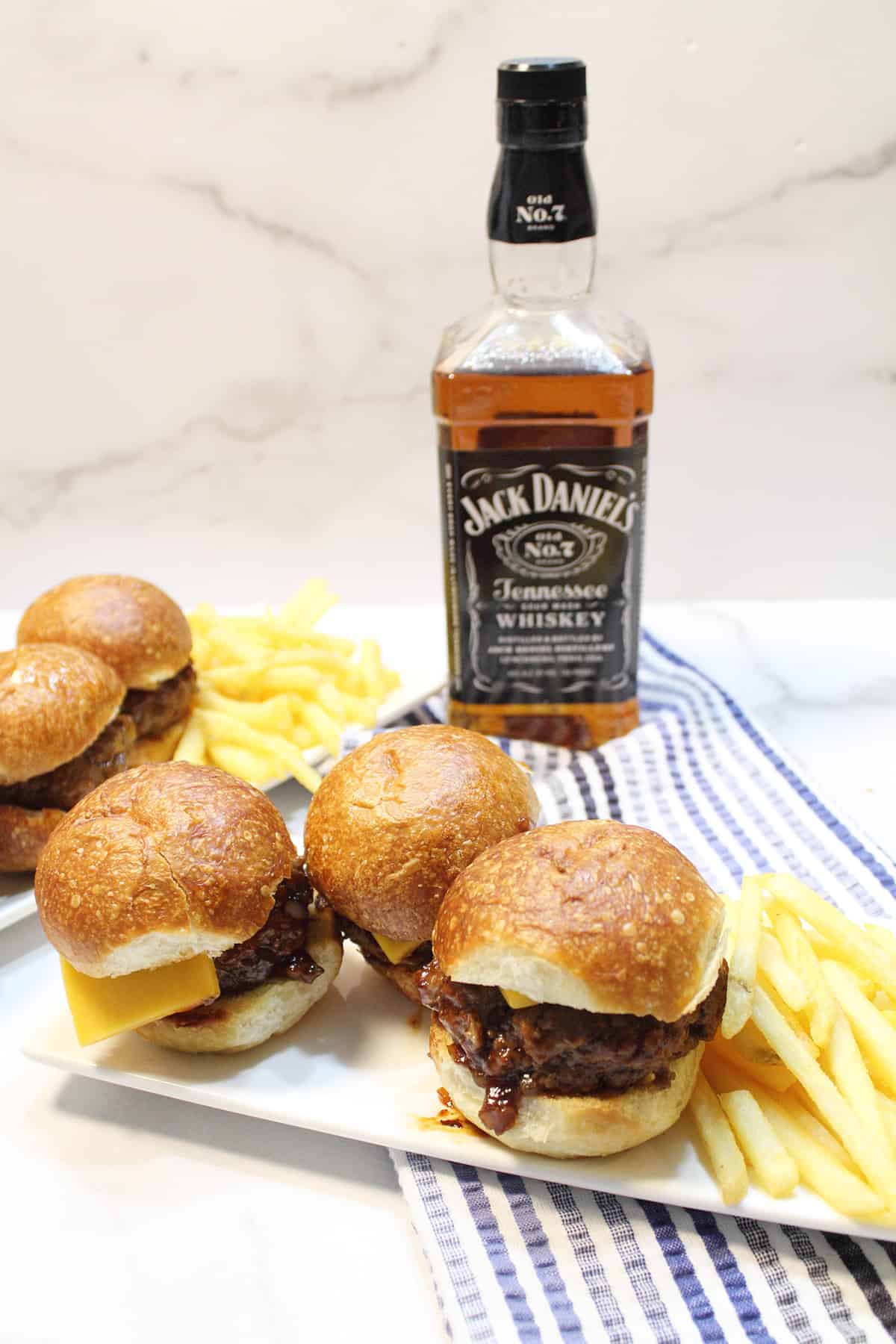 3 homemade sliders burgers on white plate with fries and whiskey bottle in background.