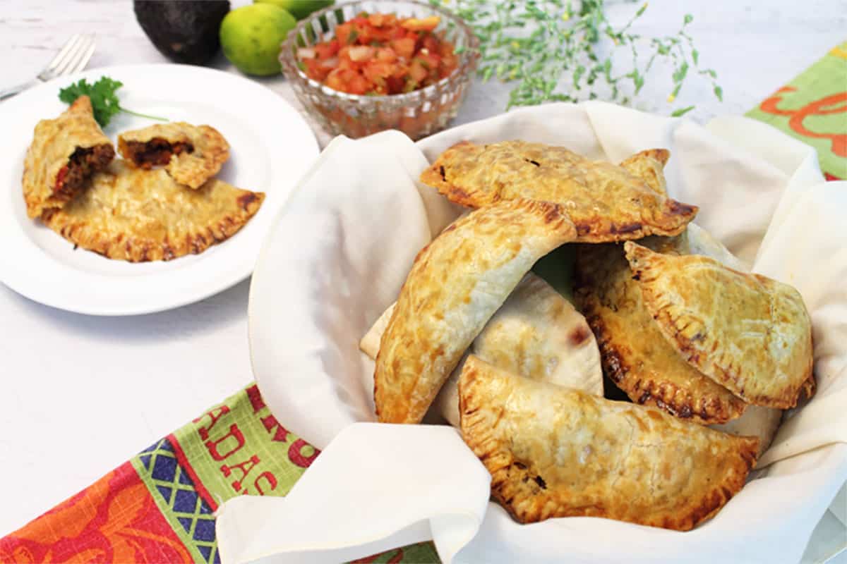 Beef empanadas in white line basket with one on plate in back.