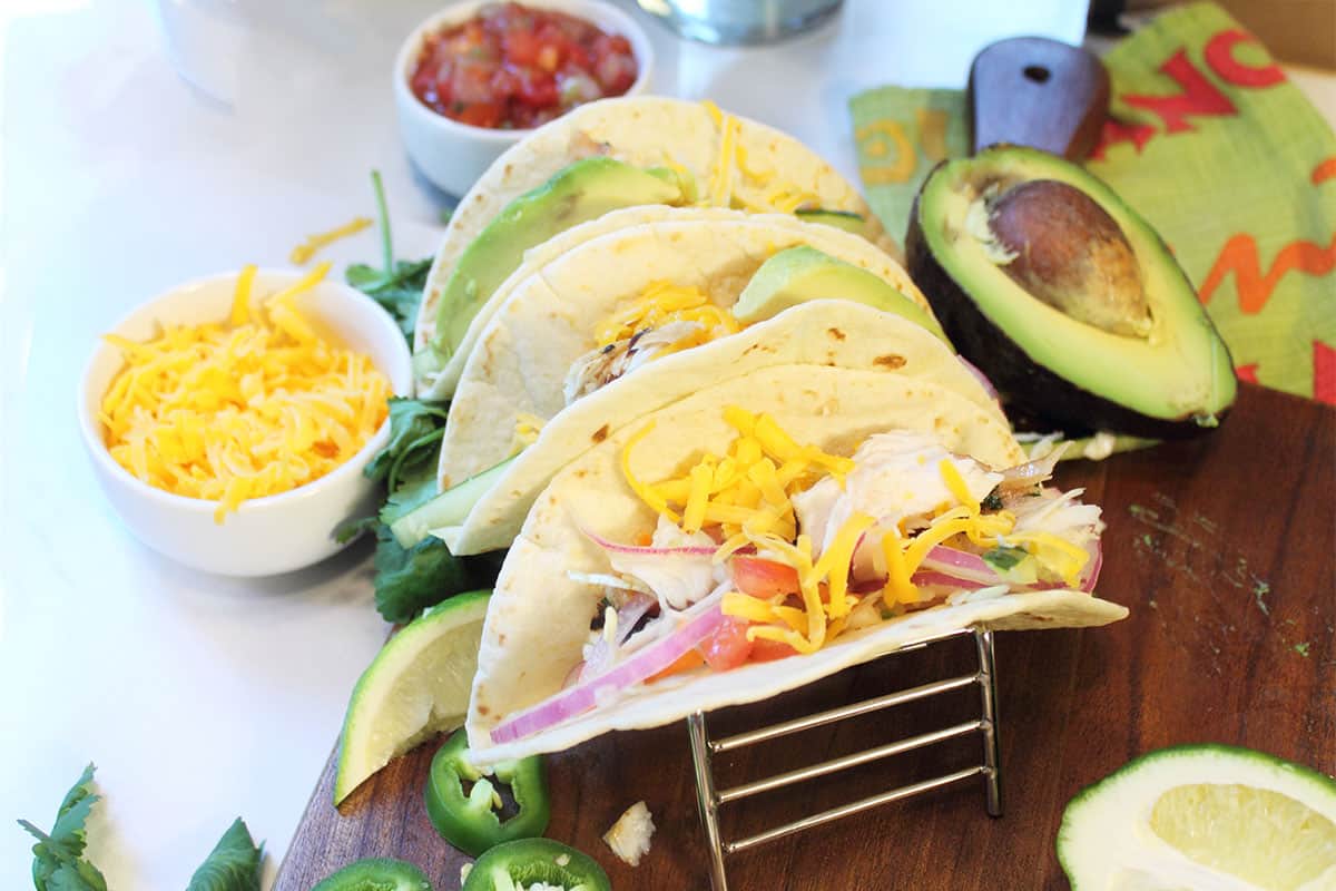 3 fish tacos in holder on wooden board with toppings around them.