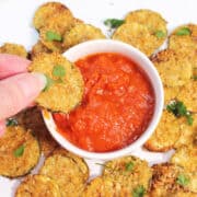 Zucchini chips surrounding a bowl of marinara and dipping one into it.