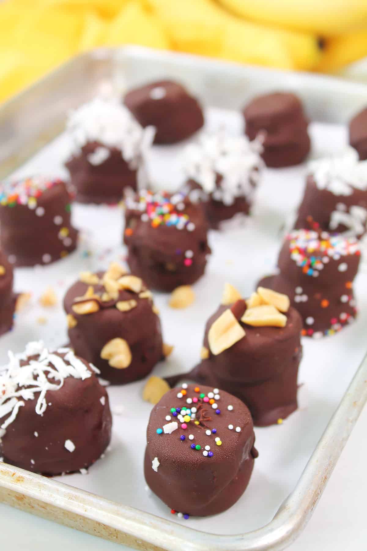 Frozen chocolate peanut butter banana bites on baking sheet with garnishes.
