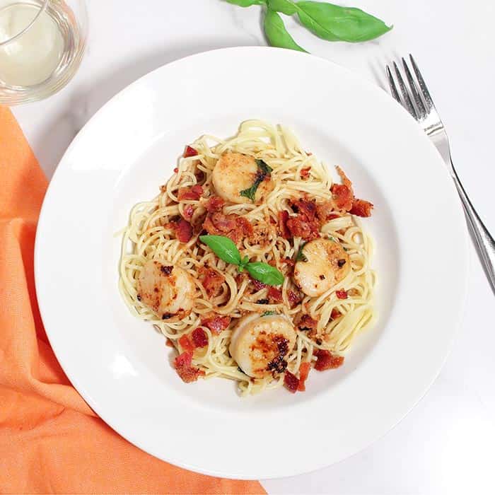 Overhead of serving of basil scallops over spaghetti in white bowl.