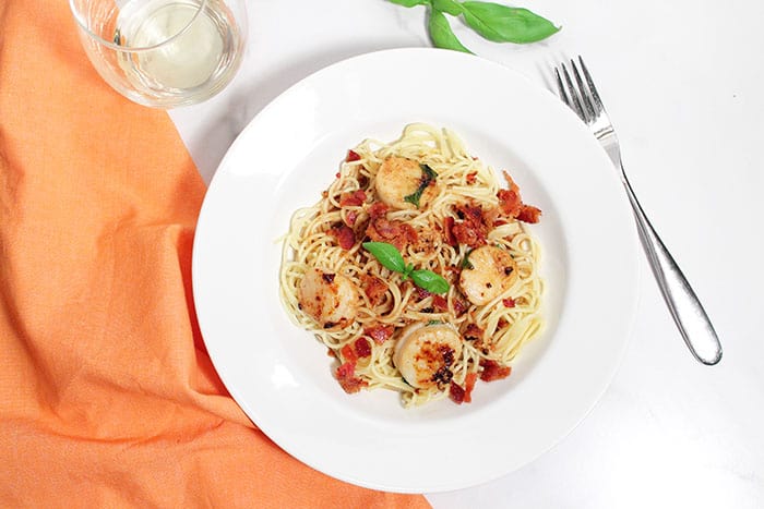 Overhead of serving of basil scallops over spaghetti in white bowl.