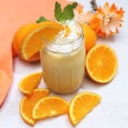 Banana Orange Smoothie with slice of orange and mint garnish and orange napkin with oranges around it.