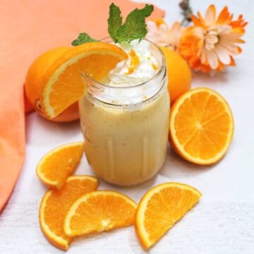 Banana Orange Smoothie with slice of orange and mint garnish and orange napkin with oranges around it.