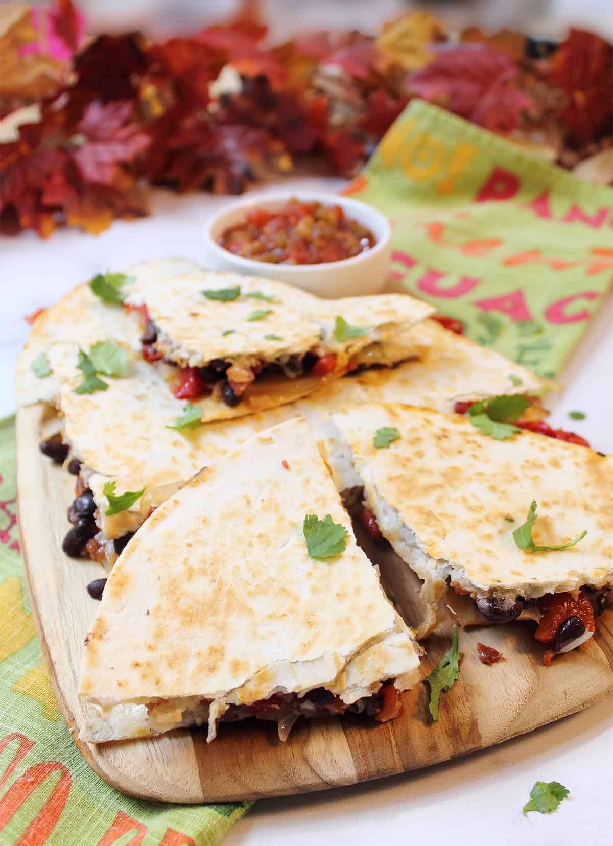 Cut roasted pepper quesadillas on board showing with salsa in back on top of Spanish worded towel.