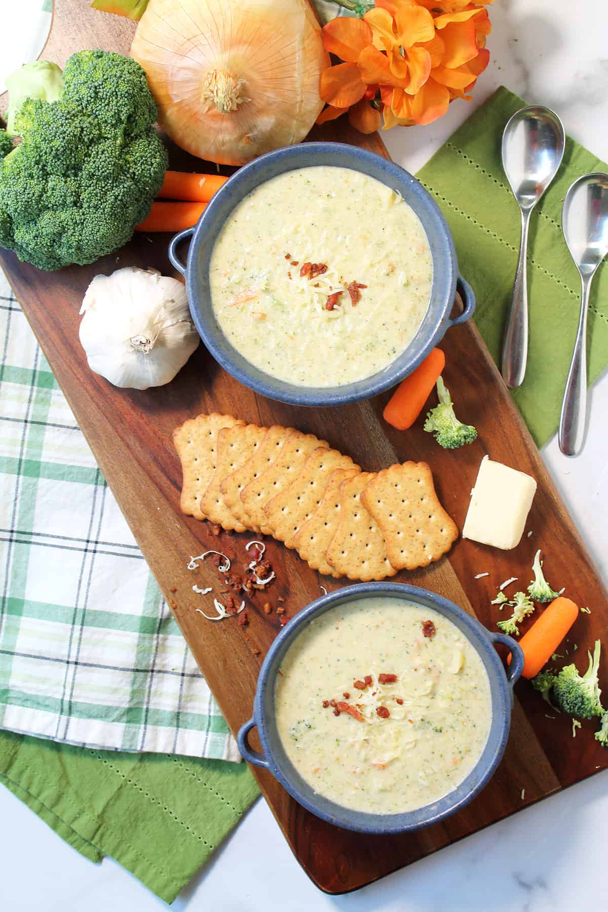 Overhead of two bowls of homemade broccoli soup on wooden board with crackers and vegetables.