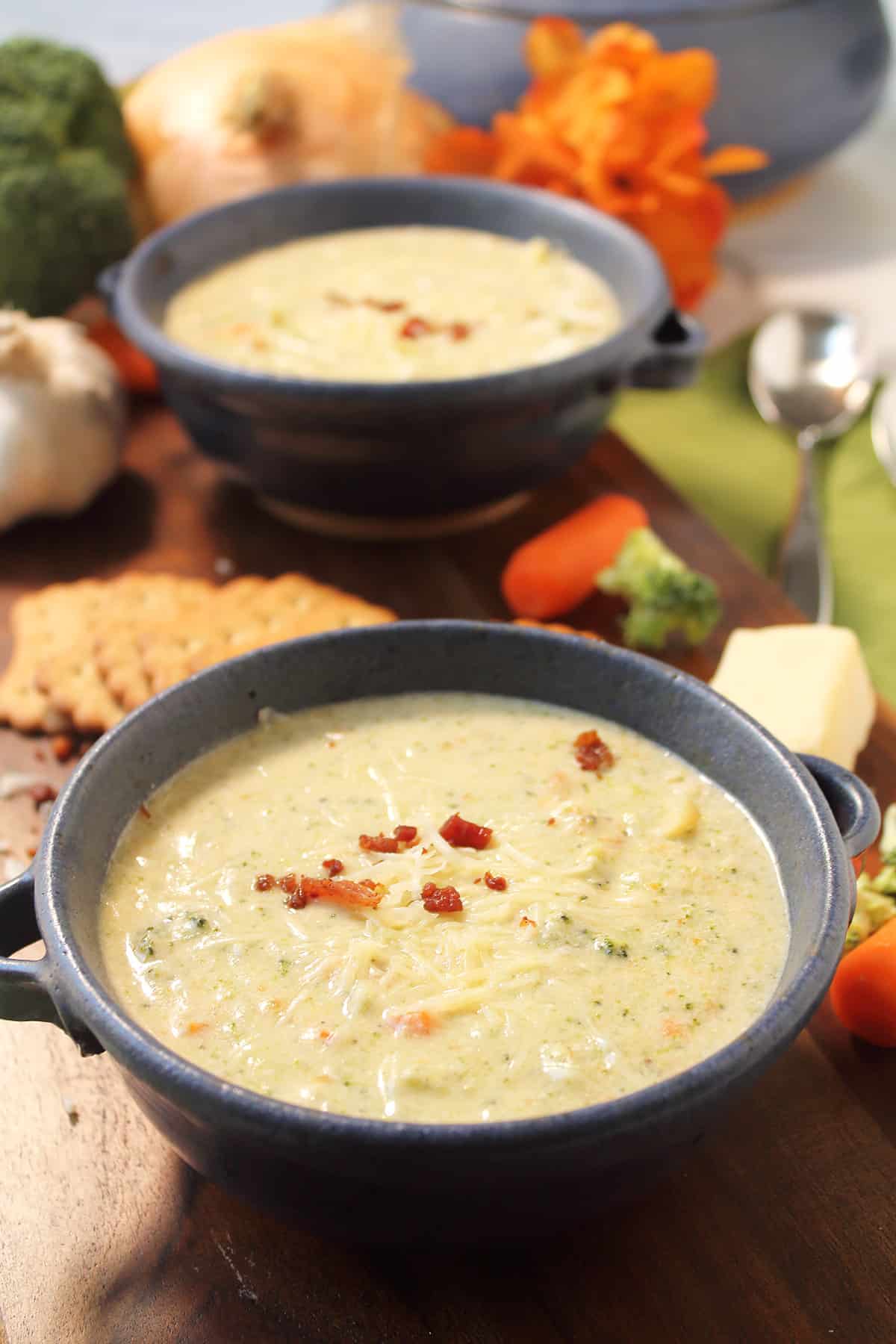 Two bowls of broccoli cheddar soup with crackers on wooden board with vegetables in back.