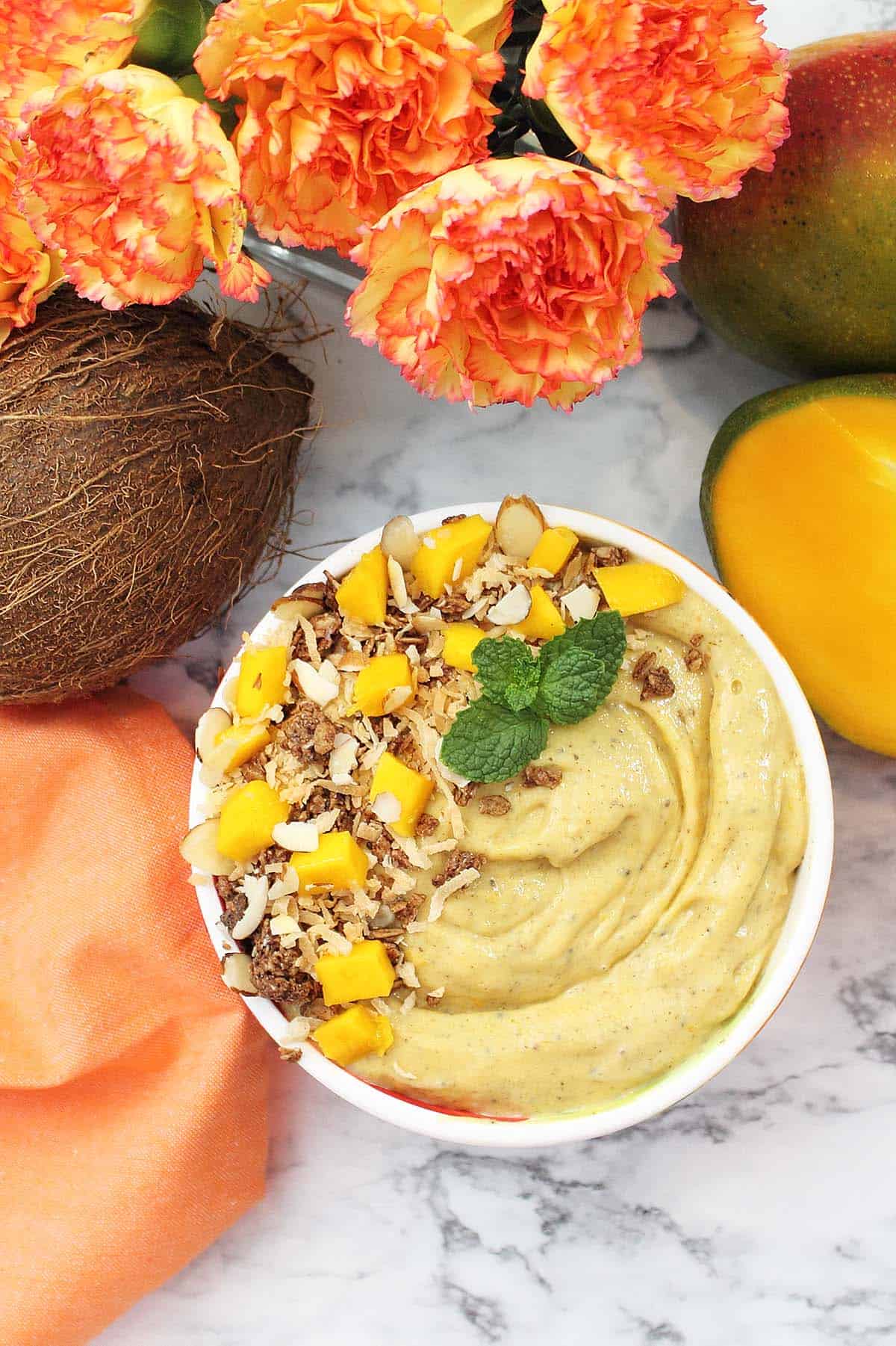 Overhead of smoothie bowl with coconut, mangoes and flowers around it