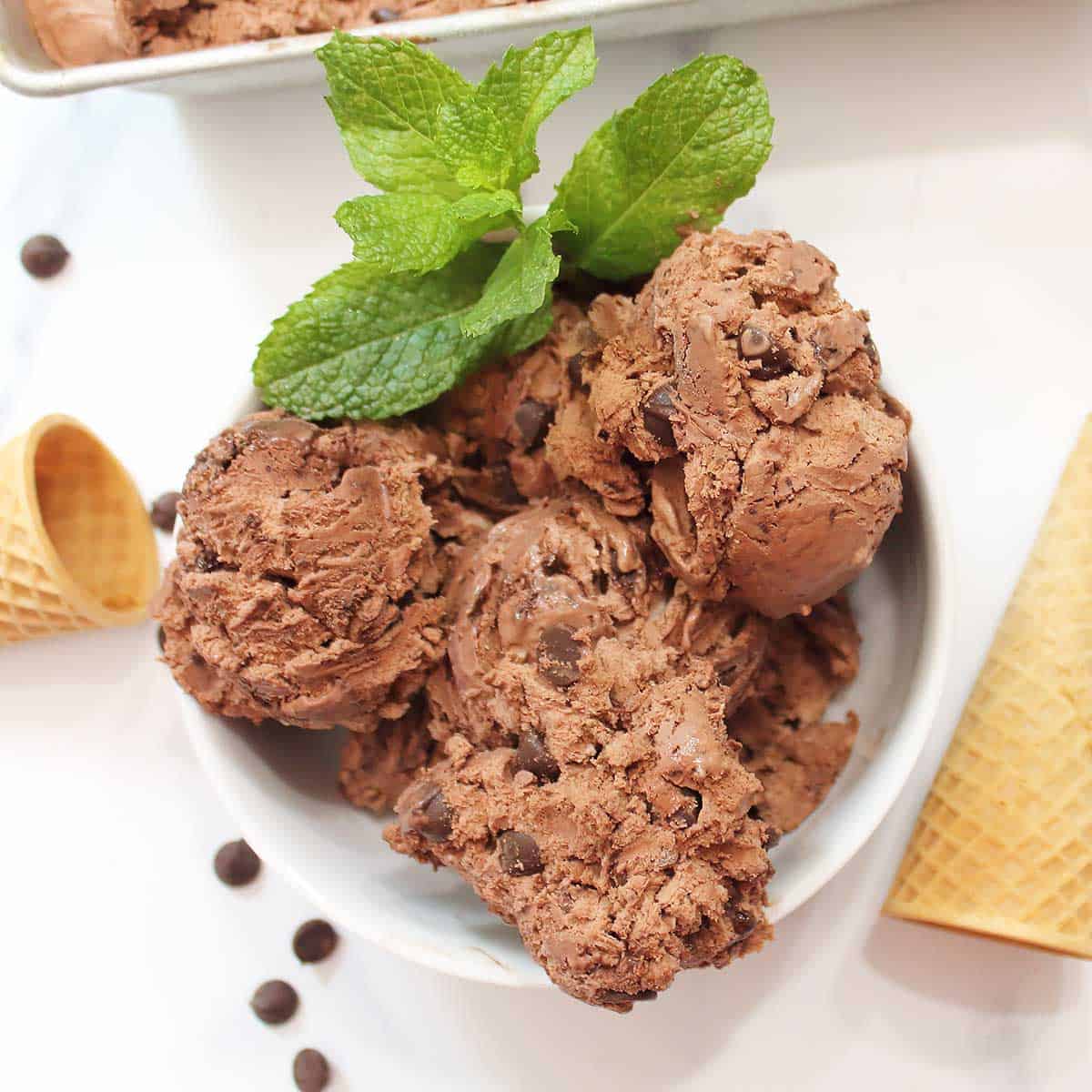 Overhead of bowl of chocolate ice cream with mint sprig and ice cream cones on white table.