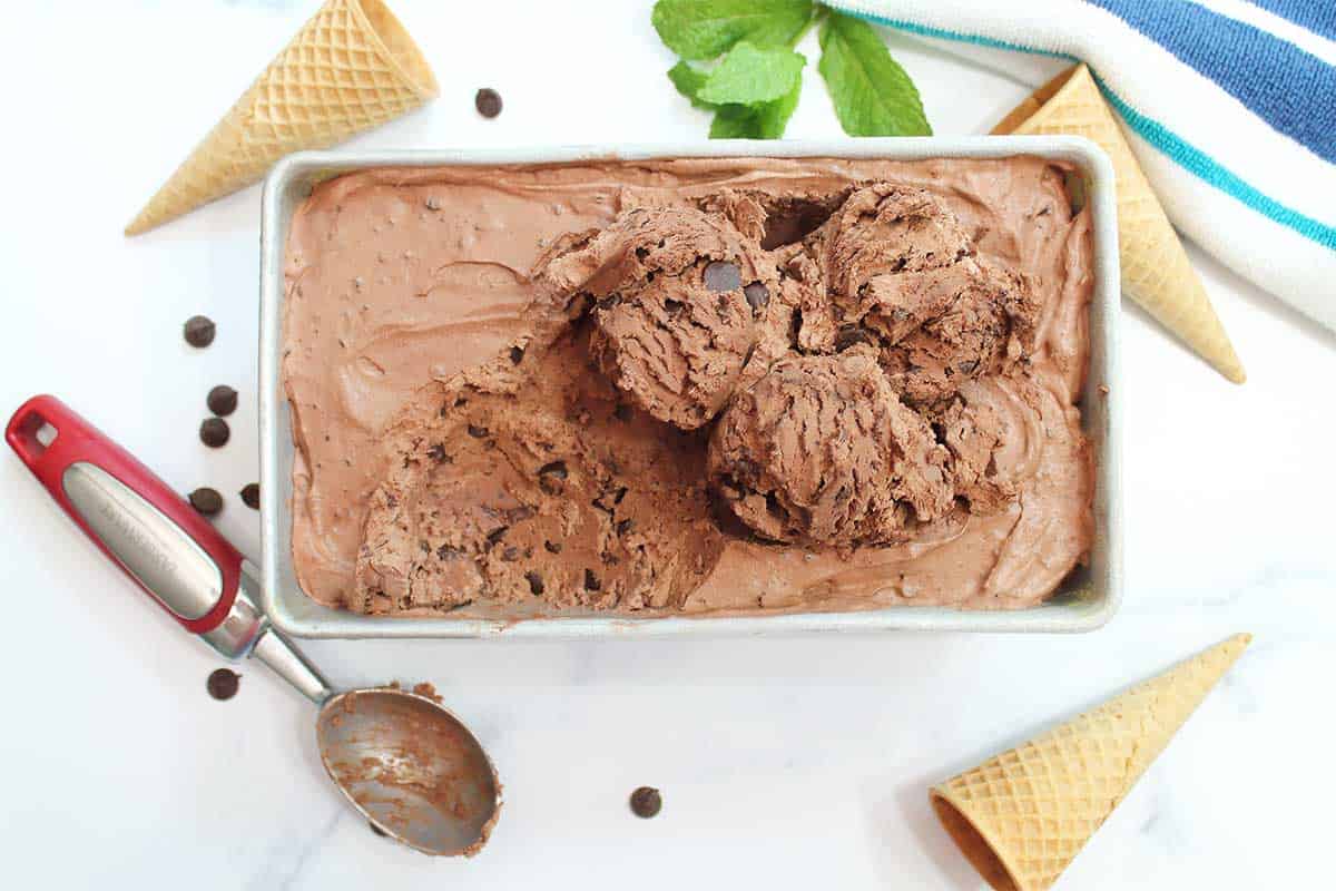 Scooped no-churn ice cream in loaf pan with ice cream scoop and sugar cones around it.