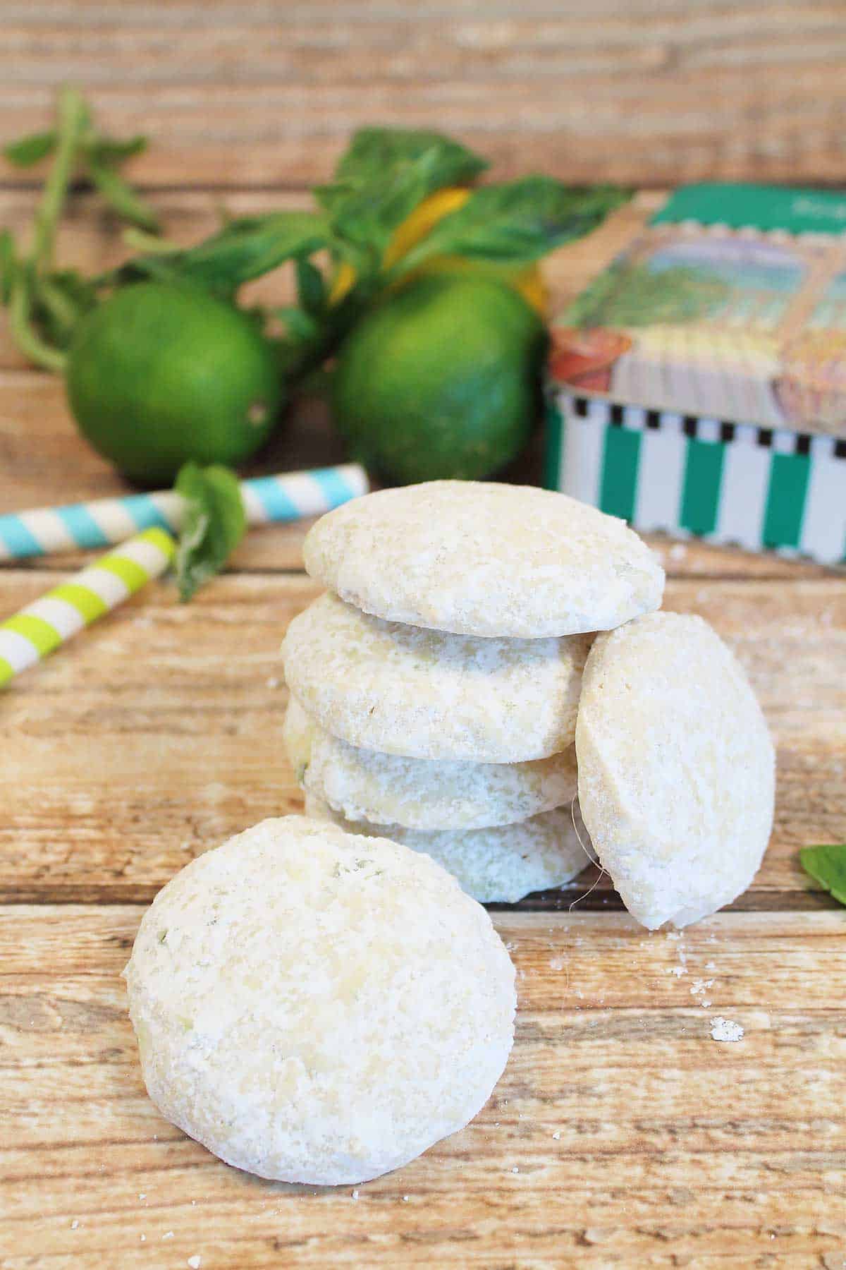 Stacked Key Lime Cookies on wooden table with limes and cookie tin in back.