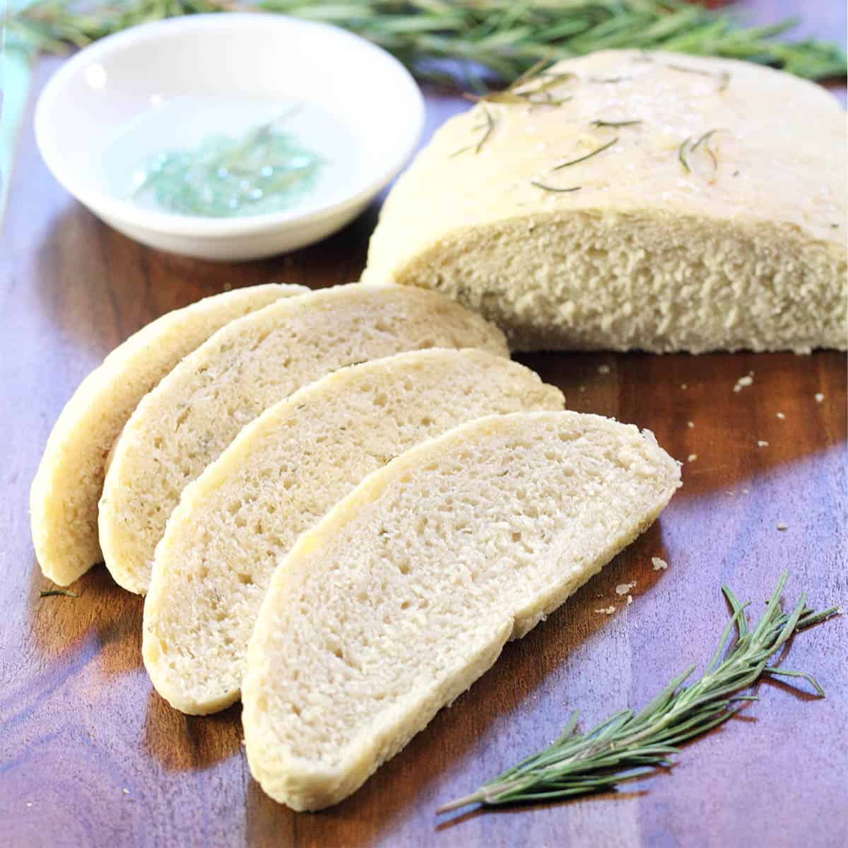 Partially sliced rosemary bread on wooden serving board with dipping bowl and rosemary.