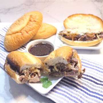 Halved sandiwch on white plate with au jus and rolls and second sandwich in back.