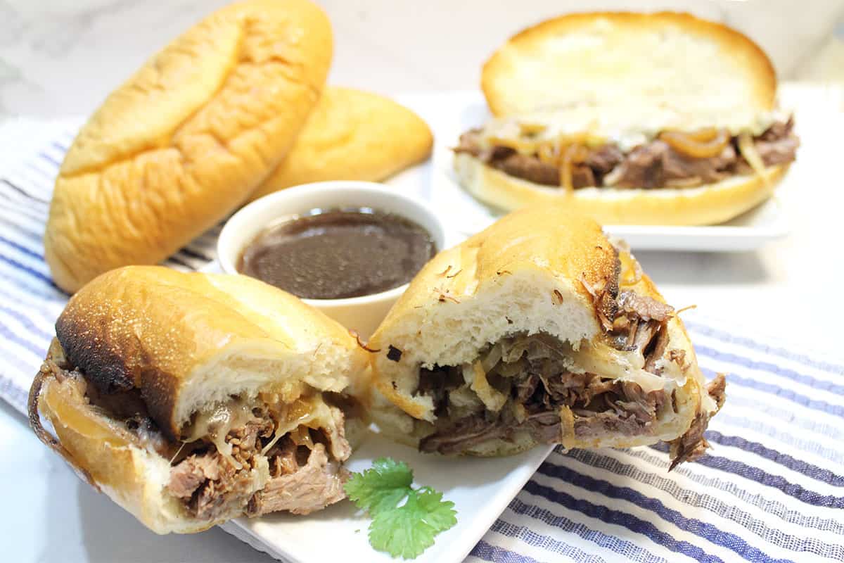 Halved sandwich on white plate with au jus and rolls and second sandwich in back.