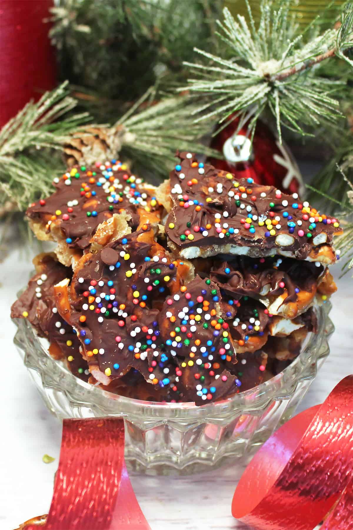 Crystal bowl of Christmas Crack with pretzels on white table with red ribbon.