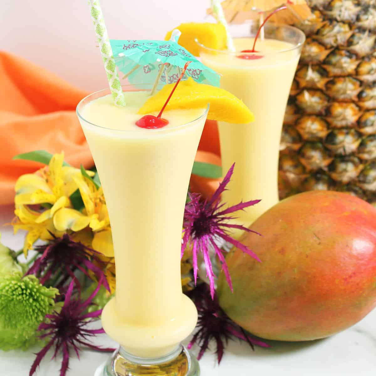 Two pina colada with mango cocktails with umbrellas near mango and pineapple.