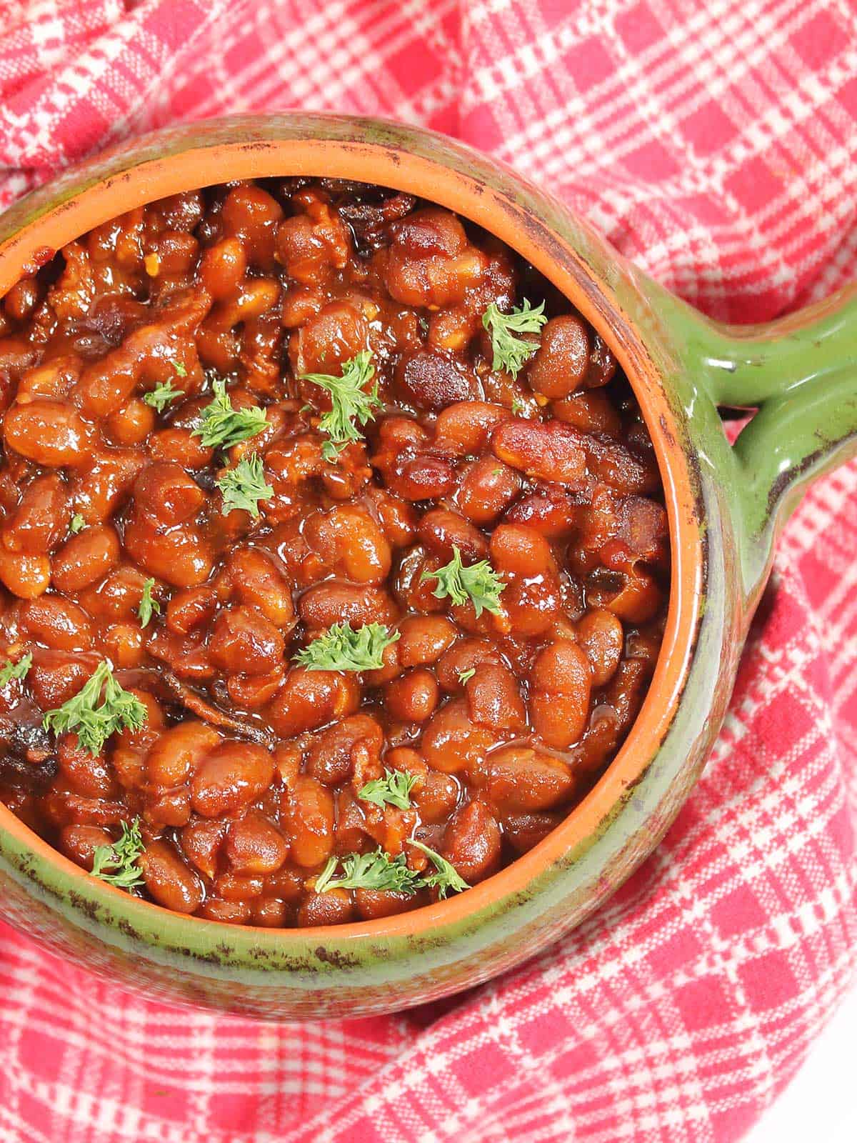 Overhead of crockpot baked beans on red checkered towel.