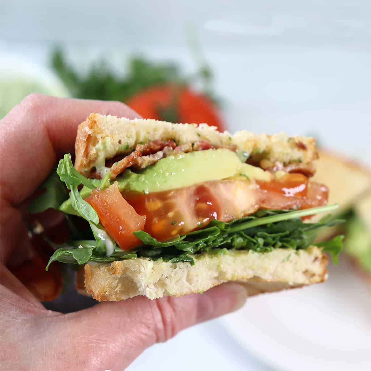 Holding halved spicy BLT sandwich with avocado.