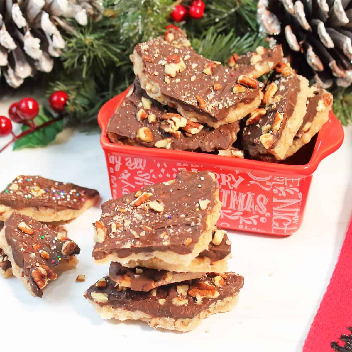 Toffee with pecans piled in front of red Christmas dish holding more toffee .