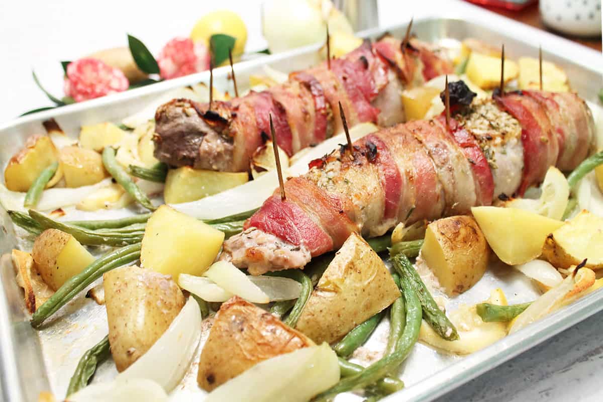 Closeup of cooked bacon-wrapped pork tenderloin and vegetables on baking sheet.