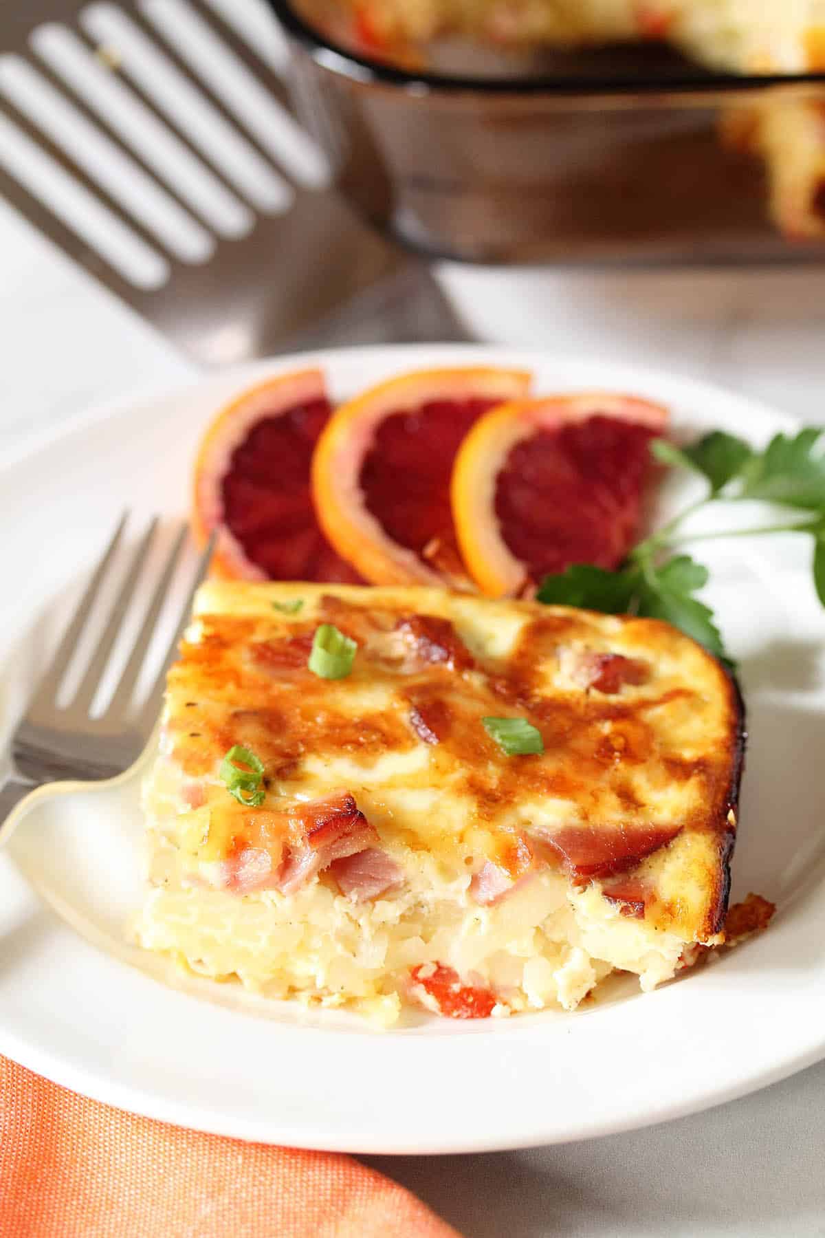 Serving of hash brown casserole with ham on white plate with blood orange slices and full casserole in back.