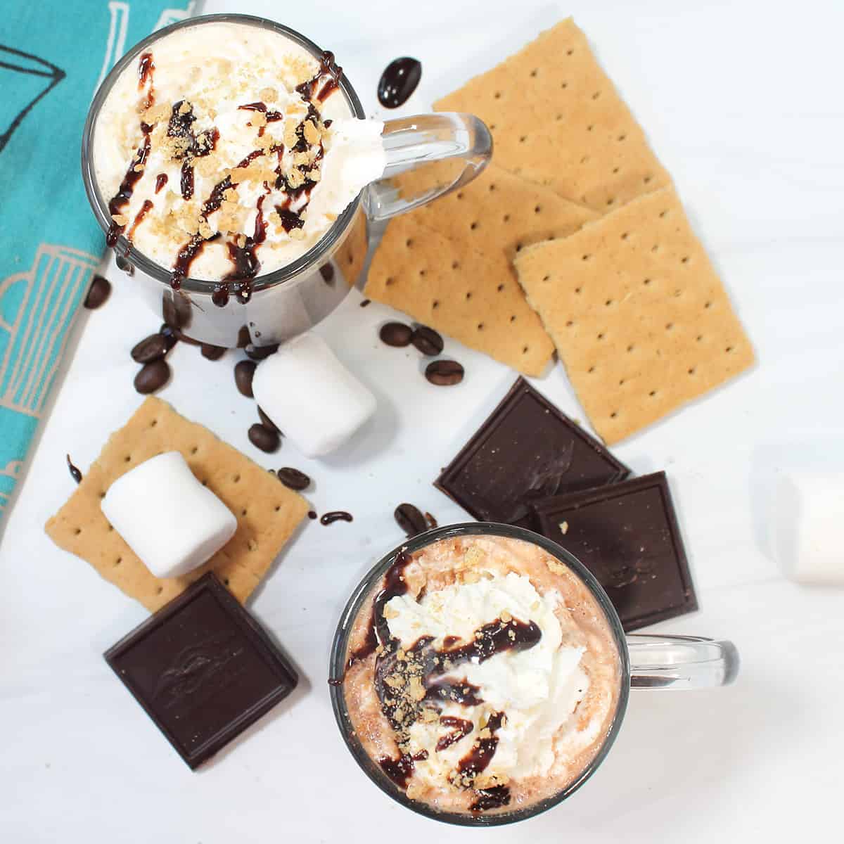 Overhead of two smores coffee with marshmallow with graham crackers, chocolate and turquoise towel.