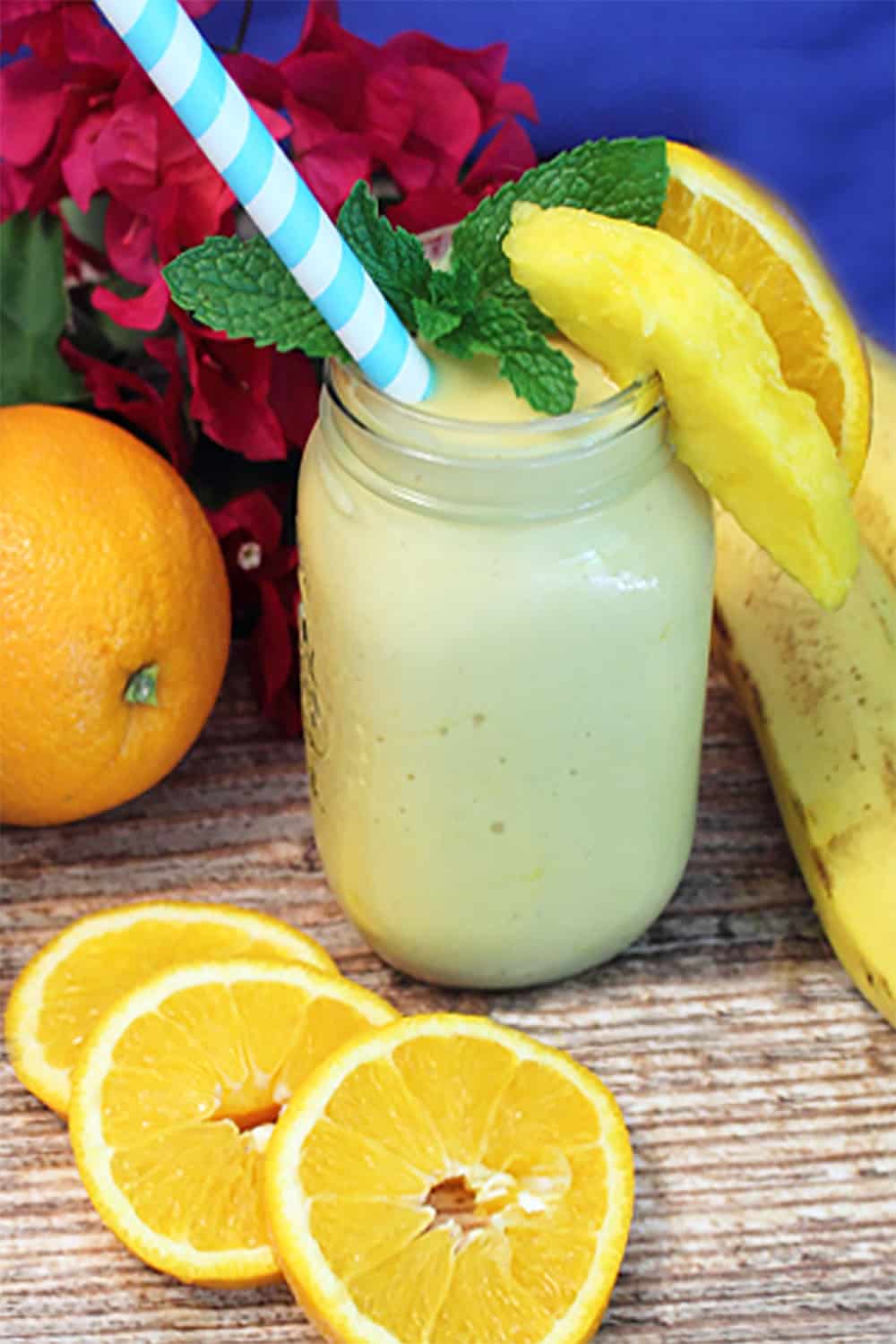 Mango smoothie surrounded by oranges bananas and mangoes on wooden table.