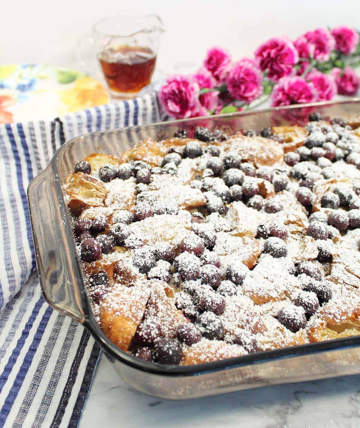 Overnight Blueberry French Toast Bake dusted with confectioner's sugar on white marble table with red flowers and maple syrup in background.
