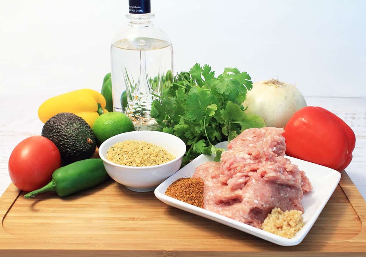 Ingredients on wooden cutting board including ground chicken onion red pepper cilantro jalapeno and tequila.