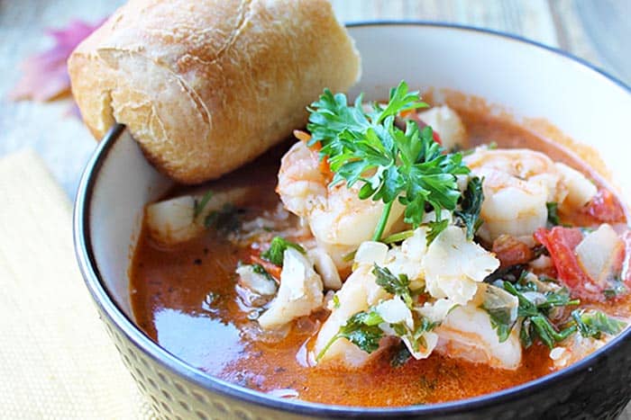 Closeup of seafood stew in brown bowl with crusty roll on side.