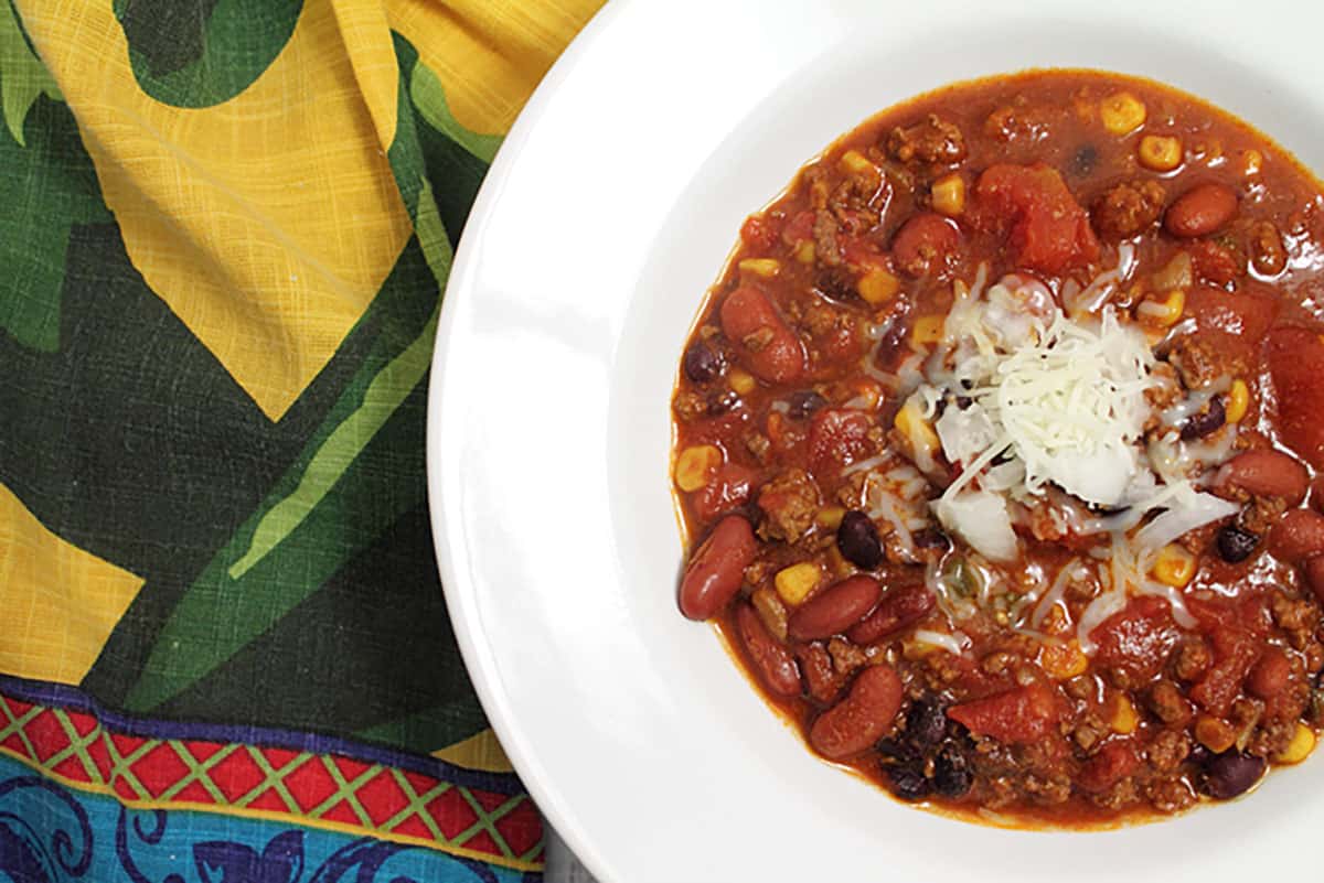 Showing partial bowl of chili in white bowl with green and gold towel beside it.
