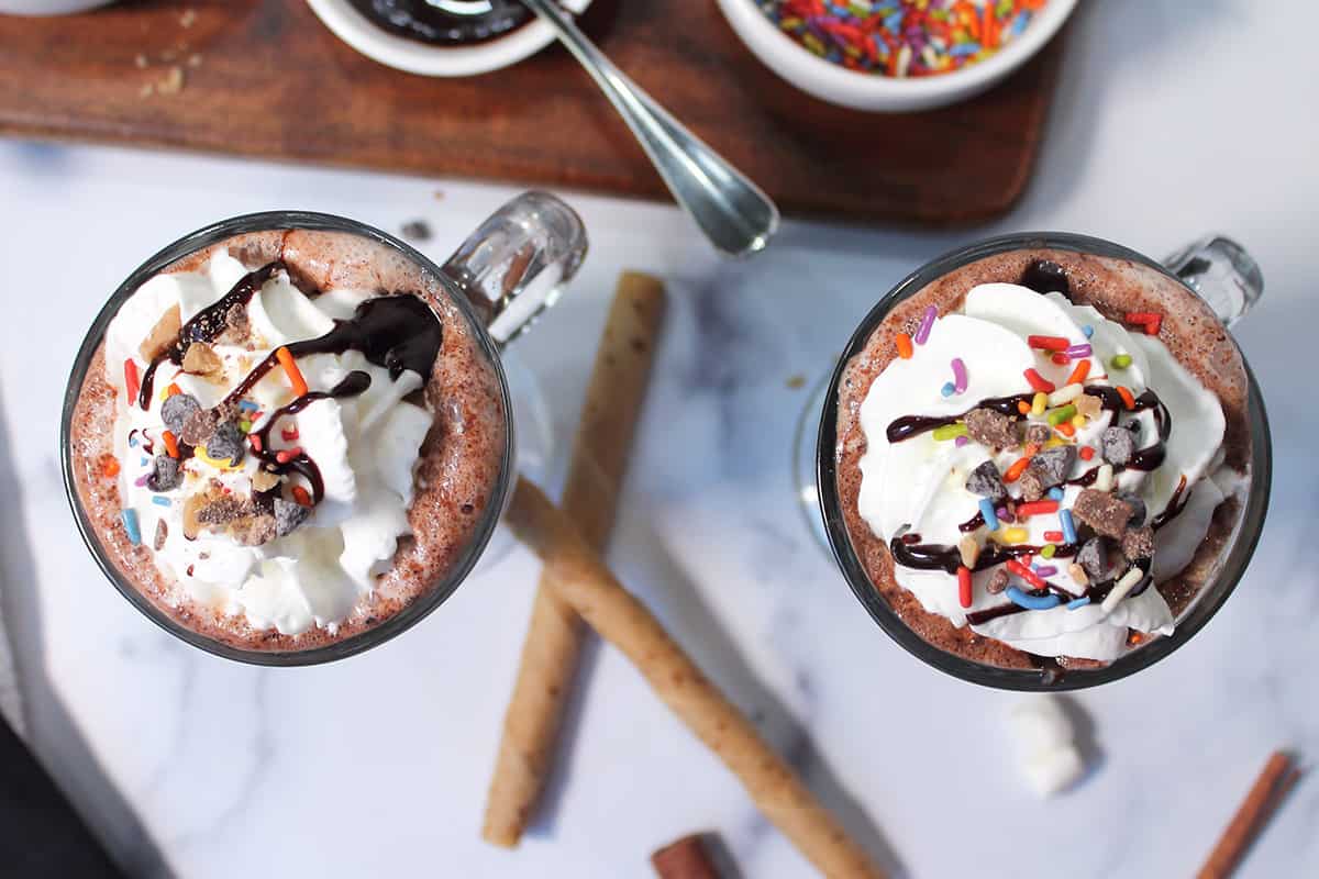 Closeup overhead of two mugs with cocoa, whipped cream and toppings.