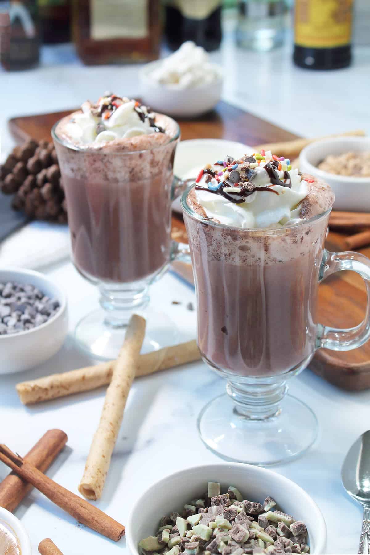 Two mugs of boozy hot chocolate with hot chocolate bar beside it and liqueurs in back.