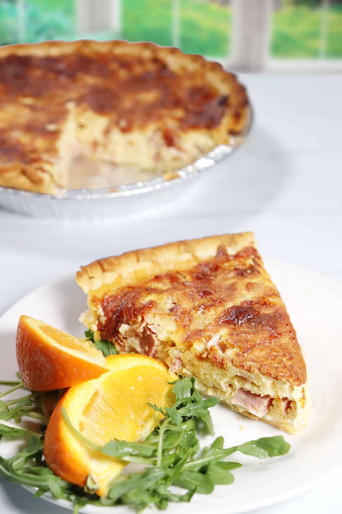 Slice of Bacon Ham and Cheese Quiche with whole pie in back.