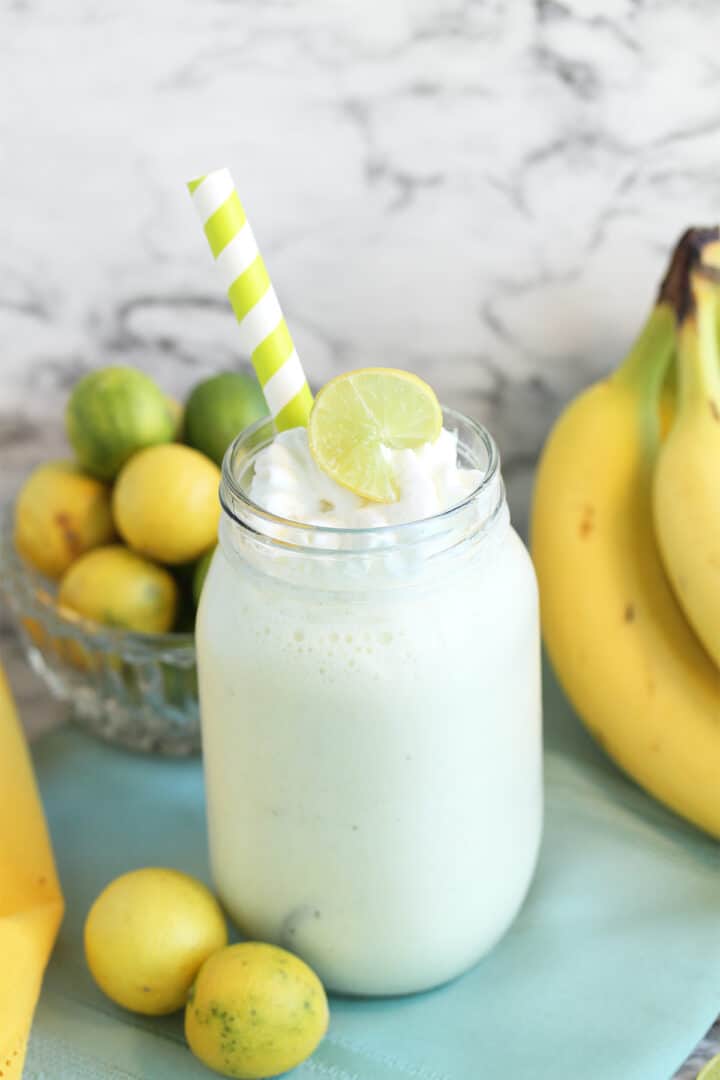 Key Lime Smoothie in mason jar with lime slice garnish and key limes and bananas behind it.