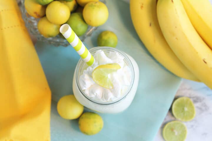 Overhead of smoothie garnished with whipped cream topping beside bananas and limes.