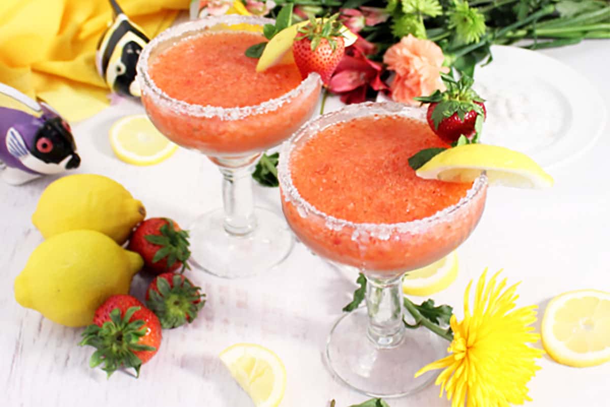 2 frozen margaritas with lemon and strawberry garnish on white table with fruit and flowers scattered around.