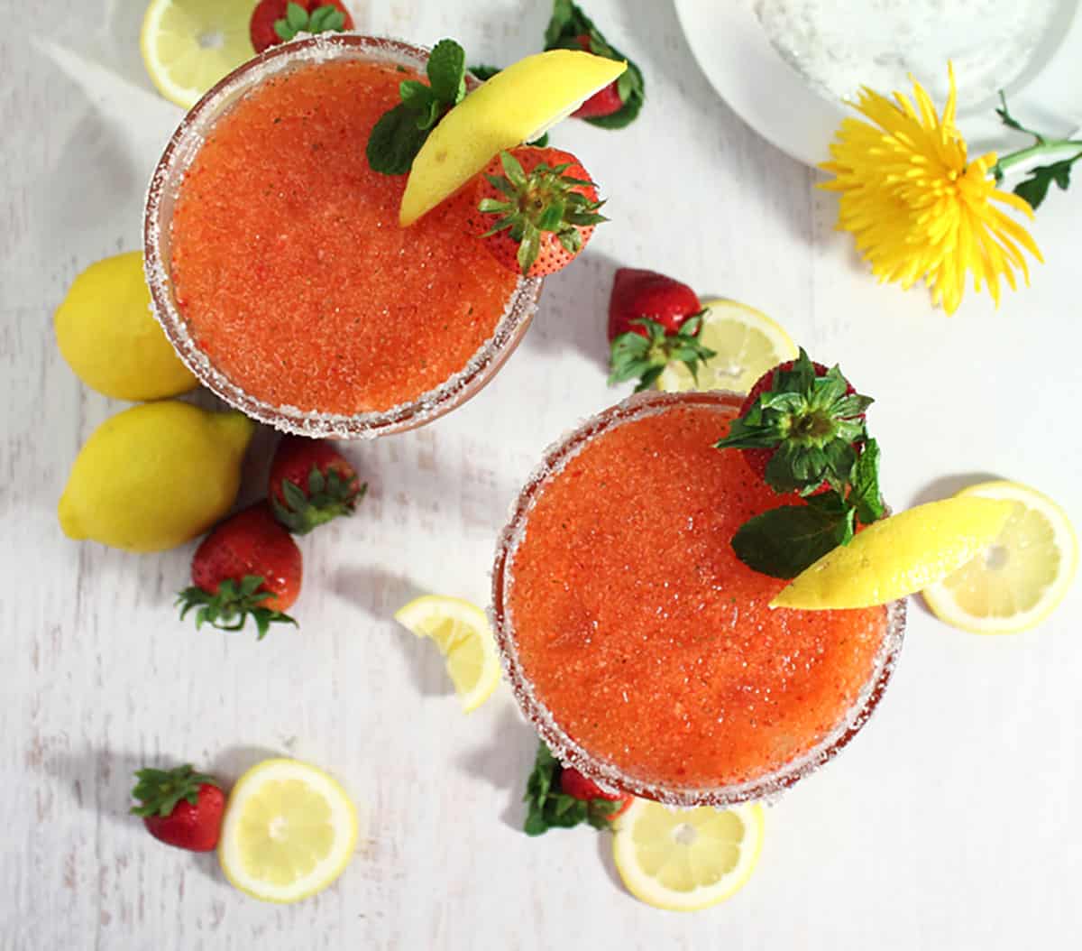Overhead of 2 cocktails with garnish of lemon wedge and strawberry on white table.