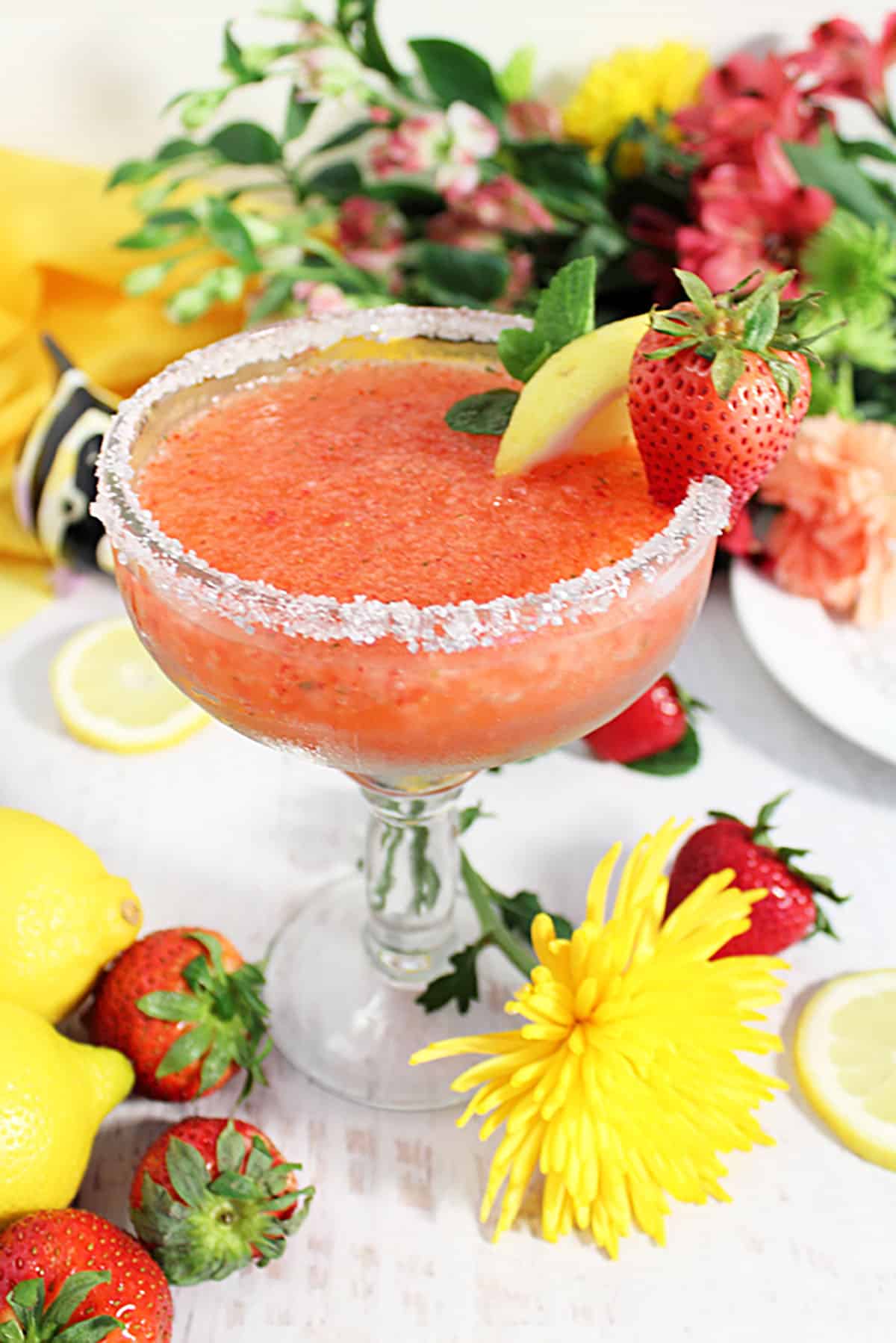 Salt rimmed glass of strawberry margarita on white table with flowers and strawberries around it.