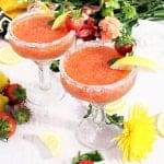 2 strawberry margaritas on white table with flowers and fruit around base.