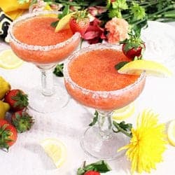 2 strawberry margaritas on white table with flowers and fruit around base.