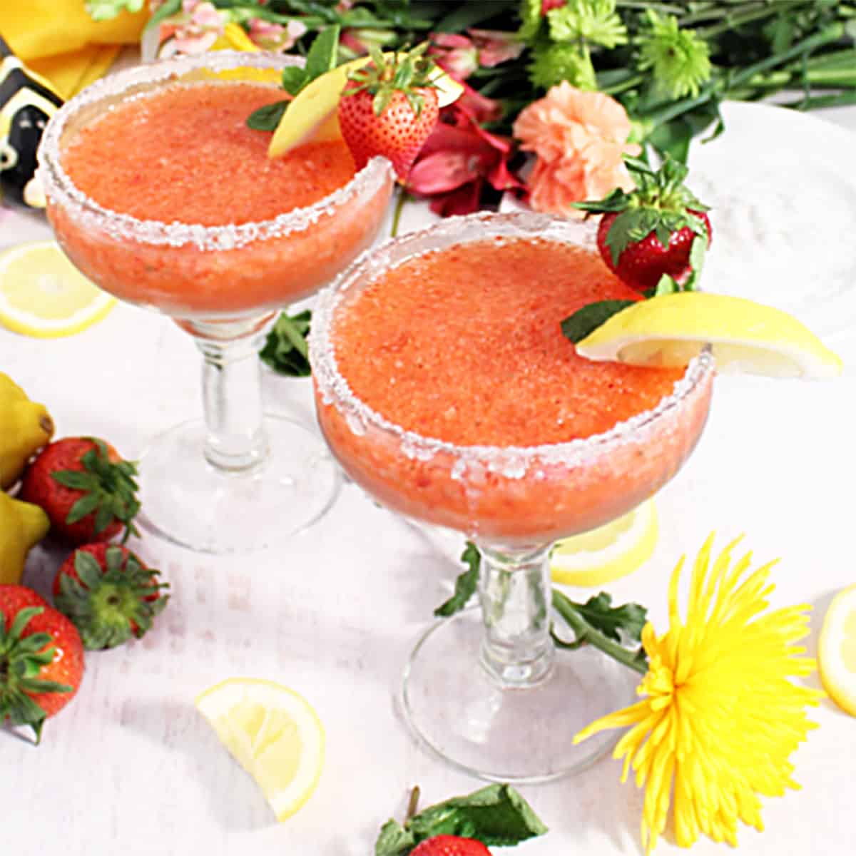 2 strawberry margaritas on white table with flowers and fruit around base.