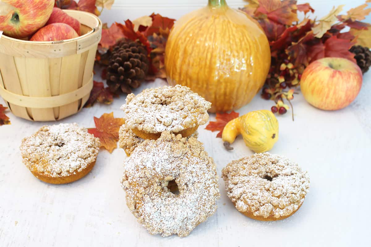 Baked donuts on white table with basket of apples and pumpkin in back.