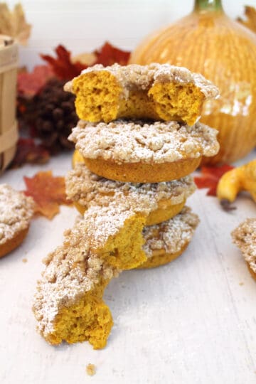 Stack of crumb donuts on white table with fall leaves and pumpkin in background.