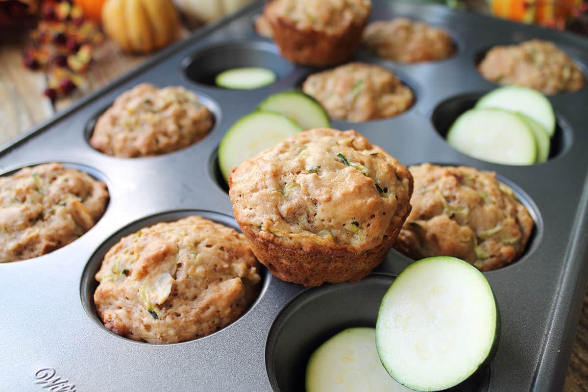 Baked zucchini muffins in muffin pan with one on top and zucchini slices in empty cavities.