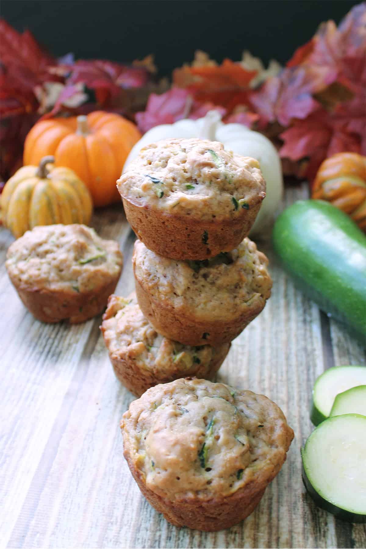 Stacked zucchini bran muffins on wooden table with fall leaves and pumpkin in back.