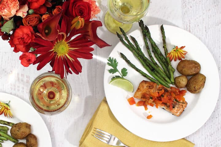 Plated grilled salmon with red pepper sauce dinner with sides of potatoes and asparagus and a glass on white wine.