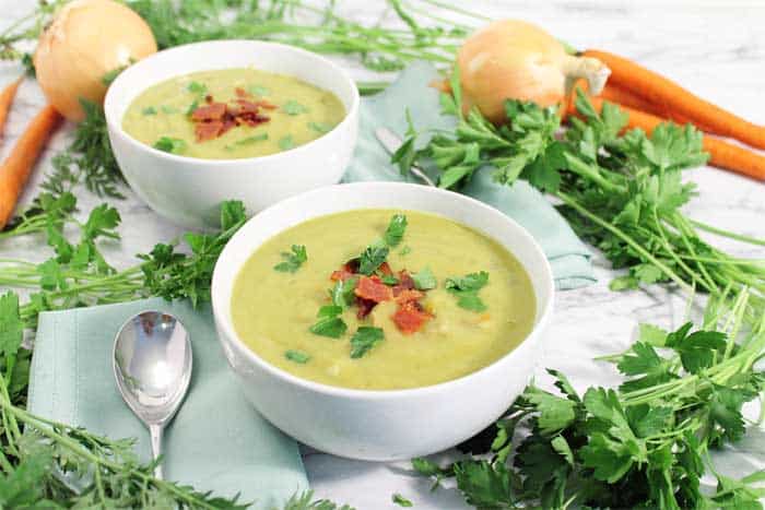 2 bowls of split pea soup on white table with greens onions and carrots around it.